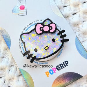 Może przedstawiać: Biało-różowy uchwyt do telefonu PopSocket Hello Kitty z błyszczącym wykończeniem. PopSocket znajduje się na białym tle z logo PopSockets i tekstem "@kawaiiicaseco" i "POPGRIP".