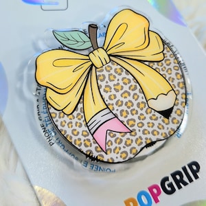 May include: A clear PopSocket phone grip with a pencil and bow design. The pencil is yellow with a pink eraser and a black tip. The bow is yellow. The background has a leopard print pattern. The word "POPGRIP" is visible.