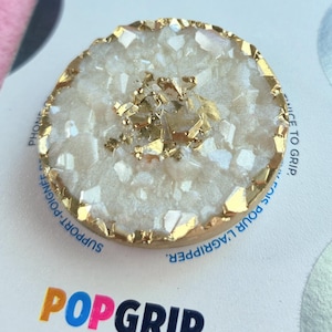Könnte beinhalten: Ein weißer und goldener PopGrip-Handyhalter mit einem kristallartigen Design. Der Halter befindet sich auf einem weißen und silbernen Hintergrund mit den aufgedruckten Wörtern "PopSockets" und "PopGrip".