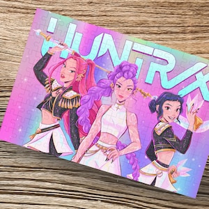 Könnte beinhalten: Ein farbenfrohes Puzzle mit drei stilisierten weiblichen Figuren in rosa, lila und schwarzen Outfits. Das Puzzle trägt das Wort "HUNTRAX" in einer Farbverlaufs-Schriftart. Eine verpackte Geschenkbox mit Bindfaden befindet sich in der oberen linken Ecke.