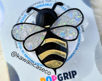 Op de afbeelding: Een zwarte en gouden bij pop grip telefoonhouder met iriserende glittervleugels. De pop grip bevindt zich op een witte achtergrond met de tekst "POPGRIP" en "GRIP. DELIX FOIS POUR L'AGRIPP." in zwart en blauw.