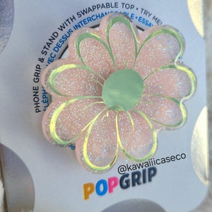 May include: A pink and green glitter phone grip with a daisy design. The grip is labeled "POPGRIP" and "@kawaiiicaseco".