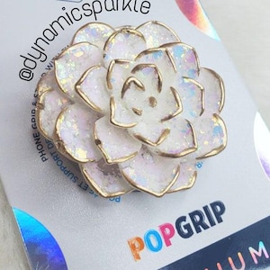 May include: A white and gold PopSockets PopGrip in the shape of a flower, with iridescent glitter accents. The PopGrip is attached to a white and silver PopSockets branded card. The card has the text "POPSOCKETS" and "POPGRIP PREMIUM".
