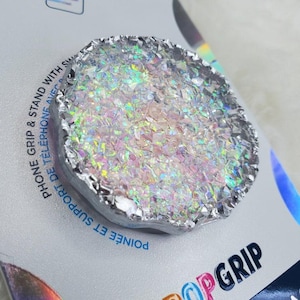 Könnte beinhalten: Ein PopSockets-Handyhalter mit einem schimmernden, irisierenden, kristallartigen Design. Das Handy-Zubehör ist rund mit einem silbernen Rand und befindet sich auf einer silber-weißen Verpackung. Auf der Verpackung stehen die Wörter "PopGrip" und "Phone Grip & Stand".