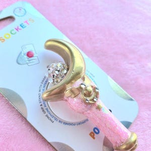 May include: A pink and gold phone grip with a crescent moon design and a sparkling crystal ball. The grip is attached to a white and blue card with the text "POPSOCKETS". The phone accessory is designed to grip and stand with swap.