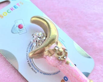 Op de afbeelding: Een roze en gouden telefoonhouder met een halve maan ontwerp en een sprankelende kristallen bol. De houder is bevestigd aan een wit en blauw kaartje met de tekst "POPSOCKETS". Telefoonaccessoire voor grip en stand.