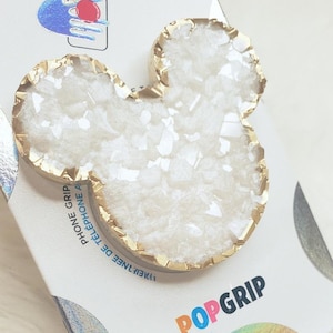 Könnte beinhalten: Ein weiß-goldenes PopGrip-Telefonzubehör in Form eines Mäusekopfes. Das Zubehör hat einen goldfarbenen Rand und eine weiße, strukturierte Mitte. Das PopGrip befindet sich auf einer weißen Karte mit dem Wort "PopGrip" in bunten Buchstaben.