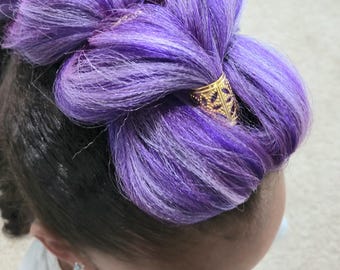 Op de afbeelding: Close-up van het haar van een kind, gestyled in een knot, met levendige paarse en witte lokken. Een goudkleurige haaraccessoire met een decoratief patroon houdt de knot vast. Het kind draagt een wit shirt en een kleine oorbel.