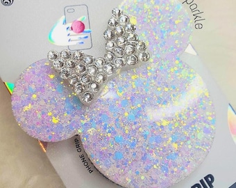 Op de afbeelding: Witte telefoon grip met een glinsterende regenboogkleurige afwerking en een zilveren strik van strass in de vorm van Mickey Mouse oren. De grip heeft de tekst "POPGRIP PREMIUM" aan de onderkant.
