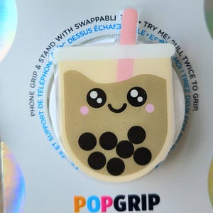May include: A PopGrip phone accessory featuring a cute boba tea design. The round, clear grip has a smiling face, pink cheeks, and black tapioca pearls. The accessory is white with the word "POPGRIP" in orange, blue, and black.
