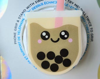Op de afbeelding: Een PopGrip telefoonaccessoire met een boba thee ontwerp. De ronde, transparante grip heeft een lachend gezicht, roze wangen en zwarte tapiocaparels. Het accessoire is wit met het woord "POPGRIP" in oranje, blauw en zwart.