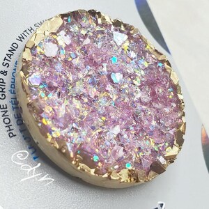 May include: A circular phone grip and stand featuring a shimmering, pale purple, crystal-like design with a gold-coloured edge. The product has the text "PHONE GRIP & STAND WITH SW" and "POPCH".