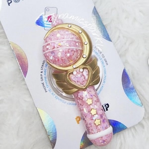 Puede incluir: Agarre para teléfono de purpurina rosa y dorada con un diseño de luna creciente y estrellas. El agarre está unido a un fondo blanco con el texto "POPSOCKETS" y "dynamicspark" impreso en él.