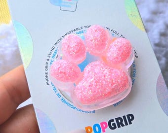 Op de afbeelding: Roze glitter telefoon grip in de vorm van een poot met de tekst "POPGRIP" op de verpakking. De verpakking bevat ook de tekst "PHONE GRIP & STAND WITH SWAPPABLE TOP".