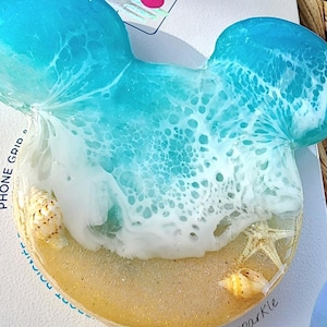 May include: A blue and white PopSocket phone grip with a beach scene. The grip features a wave with white foam, a sandy beach, and seashells. The text "POPGRIP" is printed on the bottom of the grip.