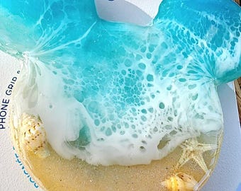 Op de afbeelding: Een blauwe en witte PopSocket-telefoonhouder met een strandscene. De houder heeft een golf met wit schuim, een zandstrand en schelpen. De tekst "POPGRIP" staat onderaan de houder.