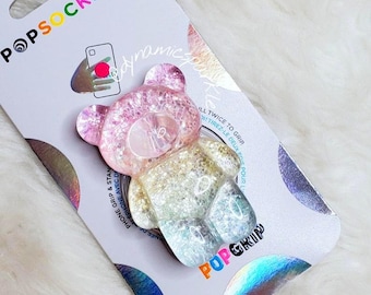 Op de afbeelding: Een telefoonhouder in de vorm van een regenboog-glitterbeer met de woorden "PopGrip" en "PopSockets" op de verpakking.