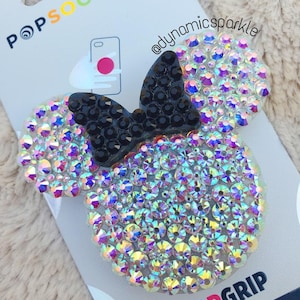 Puede incluir: Un agarre para teléfono PopSocket transparente e iridiscente con un lazo negro, con el texto "POPGRIP PREMIUM" en el empaque.