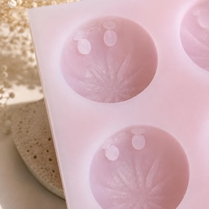 Puede incluir: Un molde de silicona de color rosa claro con múltiples cavidades circulares. Cada cavidad tiene un patrón de estrella en relieve en la base y dos pequeñas hendiduras circulares cerca del borde superior, diseñado para crear objetos detallados.