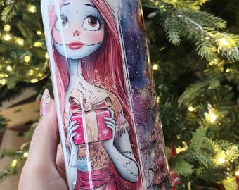 Op de afbeelding: Een roestvrijstalen beker met een kleurrijke illustratie van een personage dat een cadeau vasthoudt. Het ontwerp bevat een kerstboom en een nachtelijke hemel. De beker heeft een zilveren rietje en een doorzichtig deksel.