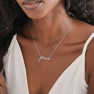 May include: A silver-toned metal necklace featuring a delicate script pendant that spells out the word Jesus. The pendant is attached to a thin, matching silver-colored chain that rests against the collarbone, creating a simple and elegant look.
