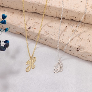 May include: Two delicate necklaces with cursive initial pendants. One is gold with the letter "H", the other is silver with the letter "B". Both necklaces hang on fine chains, displayed against a neutral background.