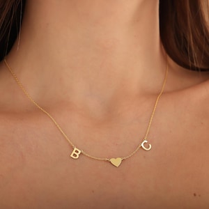 May include: A gold-toned necklace featuring a delicate chain with three suspended charms: a capital letter B, a solid heart shape in the center, and a capital letter C. The metal components have a polished finish and are evenly spaced along the chain.