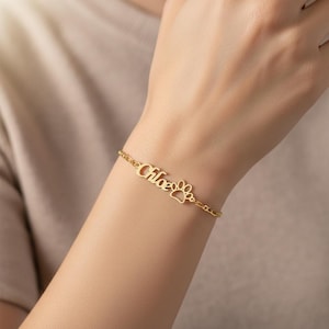 Può includere: Bracciale dorato con il nome "Chloe" e un ciondolo a forma di zampa. Il delicato bracciale a catena è mostrato su un polso, mettendo in risalto i gioielli personalizzati.