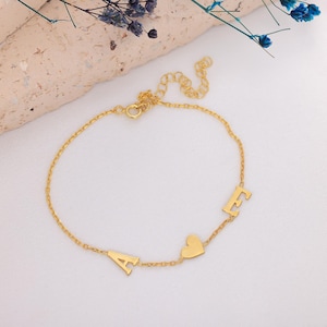Op de afbeelding: Goudkleurige armband met de letters "A" en "F", en een hartvormige bedel. De delicate kettingarmband wordt getoond op een witte ondergrond met een lichte achtergrond en enkele blauwe bloemen.