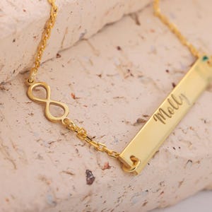 May include: A gold necklace featuring an infinity symbol and a rectangular pendant engraved with the name "Molly". The necklace is displayed on a textured, light-coloured surface. The chain and pendant are made of shiny metal.
