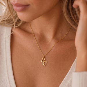 May include: A gold-toned necklace featuring two small, capital letter pendants, B and C, suspended from a delicate chain. The letters are positioned slightly overlapping each other, creating a layered effect on the thin, polished metal chain.