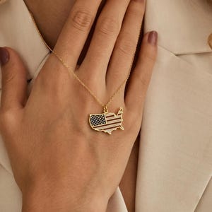 May include: A gold-toned necklace featuring a pendant shaped like the map of the United States. The pendant is filled with an enamel design depicting the American flag, showcasing a blue field with white stars and horizontal red and white stripes.