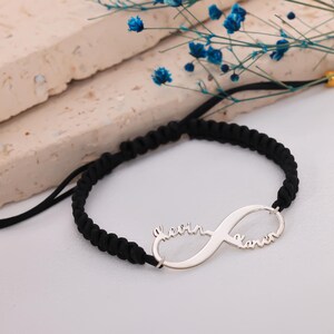 Könnte beinhalten: Ein schwarzes geflochtenes Armband mit einem silbernen Unendlichkeitszeichen. Das Unendlichkeitszeichen trägt die Namen "Kevin" und "Karen" in Schreibschrift. Das Armband liegt auf einer weißen Oberfläche mit blauen Blumen im Hintergrund.