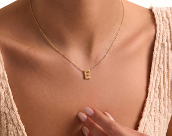 Peut inclure: Un collier doré composé d'une chaîne fine et d'un pendentif en forme de lettre E majuscule. Le pendentif est centré sur la chaîne qui repose sur la clavicule. Le métal présente une finition lisse et polie qui reflète la lumière.