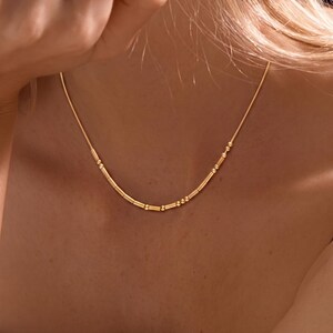May include: A gold-toned necklace featuring a delicate chain adorned with a series of small, cylindrical beads and tiny round spheres arranged in a pattern that resembles Morse code, creating a subtle and refined piece of jewelry.