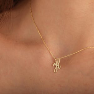 May include: A gold necklace with a cursive letter "H" pendant. The pendant is suspended from a fine chain, offering a minimalist and elegant appearance. The necklace is displayed against a skin tone background.
