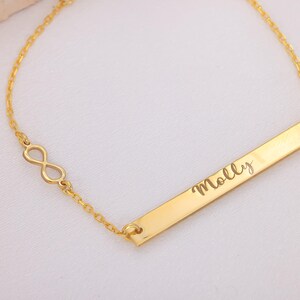 May include: A gold-colored bracelet featuring an infinity symbol and a rectangular bar. The bar is engraved with the name "Molly" in a cursive font. The bracelet is on a white background.
