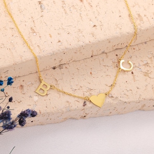 May include: A gold-colored necklace featuring the letters "B" and "C", and a heart-shaped pendant. The necklace is displayed on a textured, light-colored surface. The chain is delicate and the letters are in a serif font.