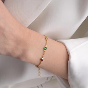 May include: A delicate gold chain bracelet featuring three small, round, bezel-set gemstones in red, green, and clear white. The bracelet includes a thin metal link chain and a short extender section with a small dangling charm at the end.