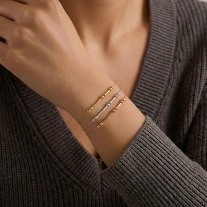 May include: Three delicate bracelets in gold, silver, and rose gold adorn a wrist. Each bracelet features a thin chain with a bar and bead design. The bracelets are displayed against a gray sweater, highlighting the jewelry's subtle elegance.