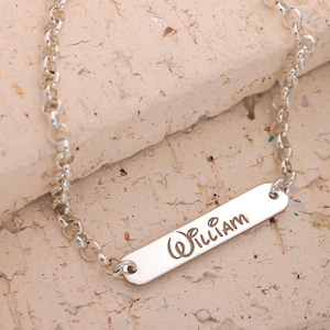 Puede incluir: Una pulsera plateada con una placa rectangular grabada con el nombre "William" en cursiva. La pulsera presenta un diseño de eslabones de cadena. El fondo es una superficie texturizada de color claro.