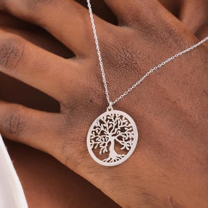 May include: A silver-toned necklace featuring a circular pendant with a detailed cutout design of a tree with spreading branches and roots. The pendant is suspended from a delicate, thin silver-toned chain that rests against the skin.