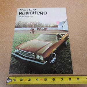 May include: A vintage advertisement for the 1973 Ford Ranchero, a pickup car. The image features a brown car parked on grass, with a white fence, trees, and a small building in the background. The text "1973 Ford Ranchero The Pickup Car" is at the top.