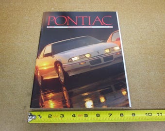 May include: A magazine cover featuring a white Pontiac car with the word "PONTIAC" in red at the top. The magazine is on a light brown surface. A yellow measuring tape is at the bottom.