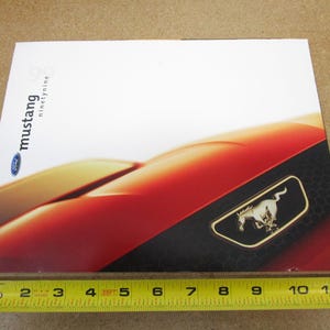 May include: A red Ford Mustang brochure with the text "Mustang ninety nine" and a close-up of the front of the car with the iconic horse logo.