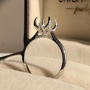 Puede incluir: Un anillo de plata con un diseño único en forma de corona, engastado con pequeñas piedras brillantes. El anillo se muestra dentro de una caja de joyería con el nombre de la marca "UNICASTON" visible. El diseño del anillo presenta un engaste central ornamentado.