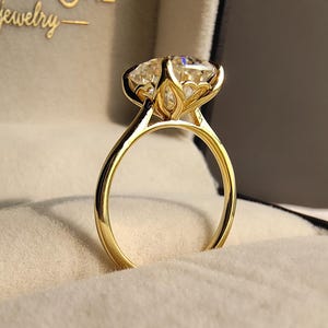 Könnte beinhalten: Ein goldener Ring mit einem großen, klaren Edelstein. Der Ring hat eine Vier-Zacken-Fassung und wird in einer Schmuckschatulle präsentiert. Der Text "UNICASTONE - jewelry" ist in goldfarbenen Buchstaben zu sehen.