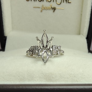 May include: A silver-colored ring with a floral design, set in a black jewelry box with a white interior. The ring features a central flower-like element and smaller stones on either side. The box has the text "UNICASTONE - jewelry" in gold.