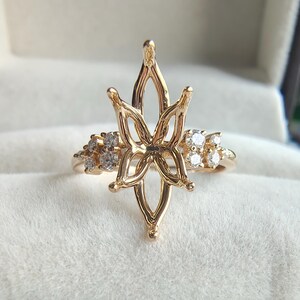 May include: A gold ring setting featuring a central marquise-shaped prong mount with a floral-inspired design, flanked by clusters of small round clear stones on each side. The text UNICASTONE jewelry is visible in the background.