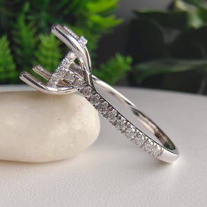May include: A silver-toned metal ring featuring a high-set prong mounting designed to hold a centre stone. The band is adorned with a row of small, round, clear pavé-set stones that extend along the shank towards the elevated setting.